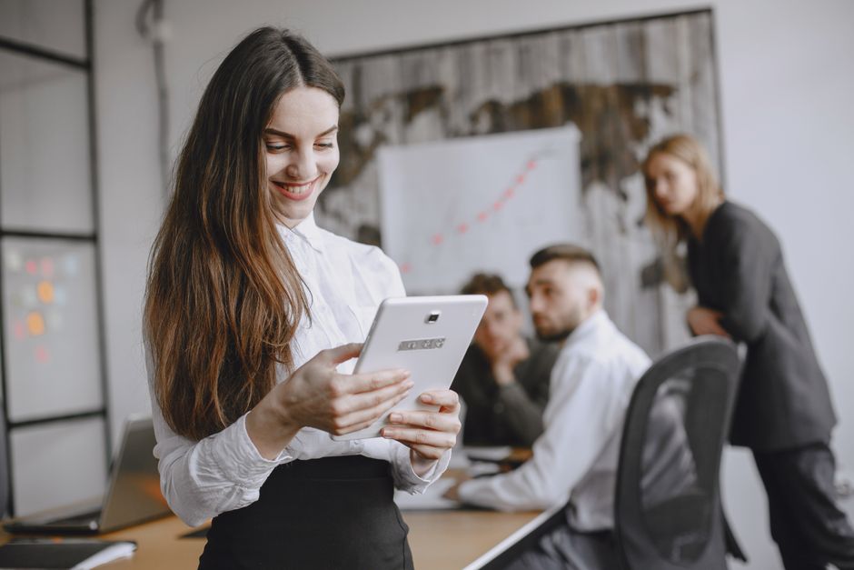 Young woman in a professional office setting browsing a tablet with colleagues in the background.